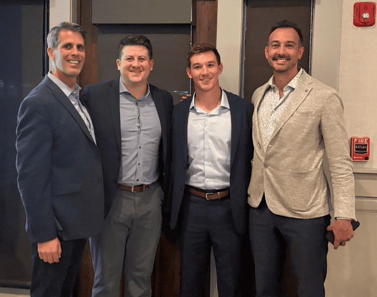 Four men in suits posing together and smiling in an indoor hallway or lobby.